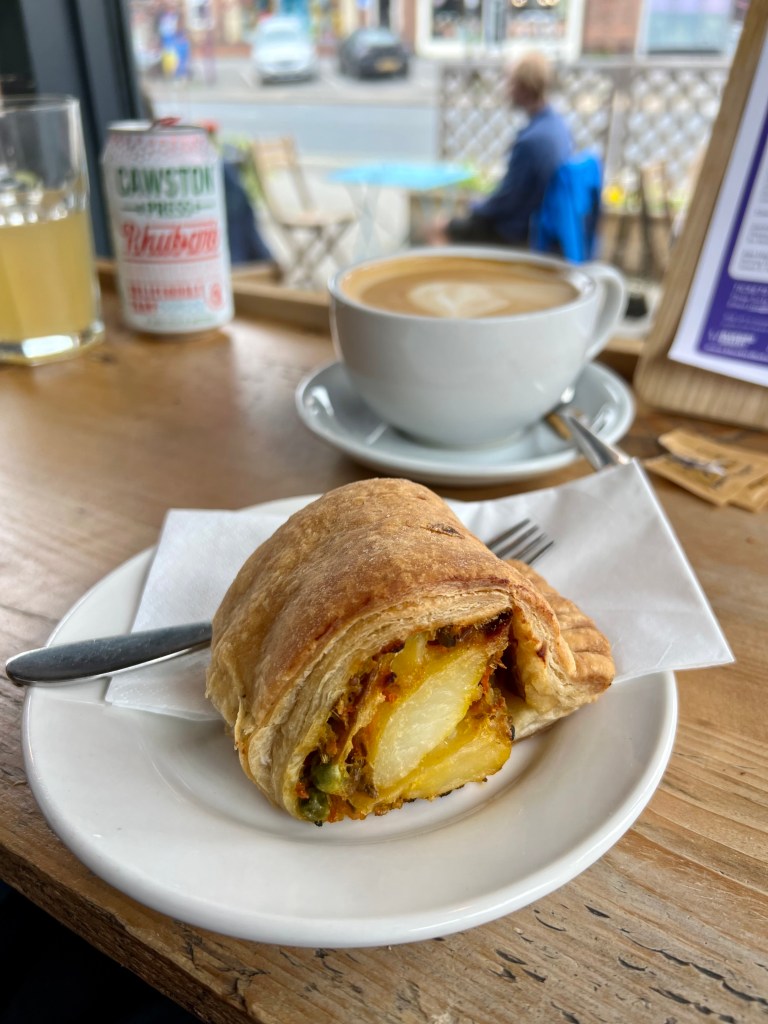 The photo shows a small white plate with a large sausage roll type pastry with a potato filling. Behind is a cup of coffee in a white cup and saucer. 