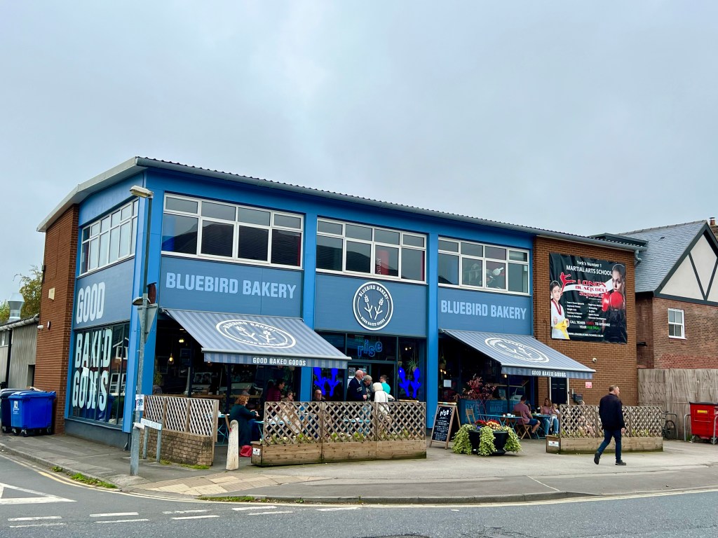 The photo is a shot of the bluebird Bakery from the other side of the road. It is a 2 storey building with a row of windows running around the second floor. The ground floor has a blue striped awning on either side of the central door and the building is painted blue. Bluebird bakery is painted in large letters above each awning and Good Baked goods on the end of the building. There is a wooden fence surrounding the outside seating areas on each side of the building. 