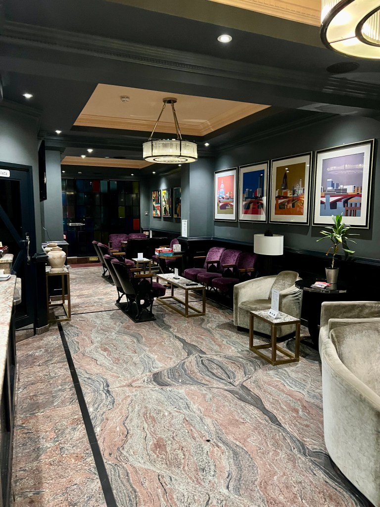 The photo shows the lounge area of The California Hotel. The floor is a coral and black swirly marble design and the walls are painted a dark green colour. At the far end of the room there are purple velvet sofas and in the foreground silver velvet armchairs. There are 2 glass circular lamp shades hanging from the ceiling.