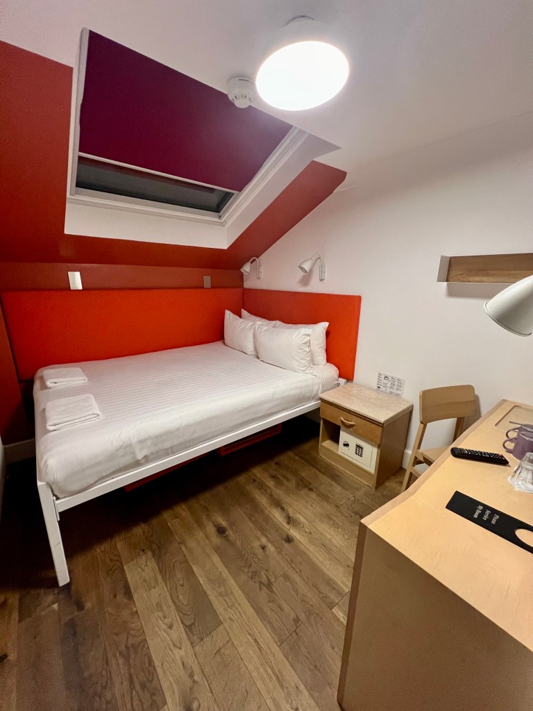 A photo of the room looking towards the double bed. The right handside of the bed is up against the back wall. The bed is under a large skylight with a red blind. The back wall is painted red and the wall on the right is painted white. There is a wooden bedside cabinet next to the bed and a wooden desk in the foreground of the photo.