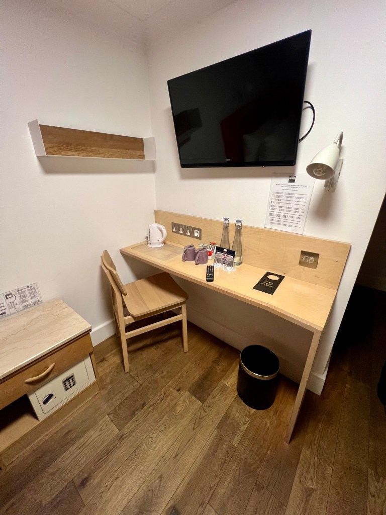 The photo shows the desk area of the room with white walls and a laminate wood floor. The desk is against the wall with a large flat screen TV on the wall above it. There is a wooden chair at the desk and a wooden bedside table on the left hand-side of the photo. On the desk there is 2 glass bottles of water, 2 purple mugs upside down, tea and coffee and a kettle.