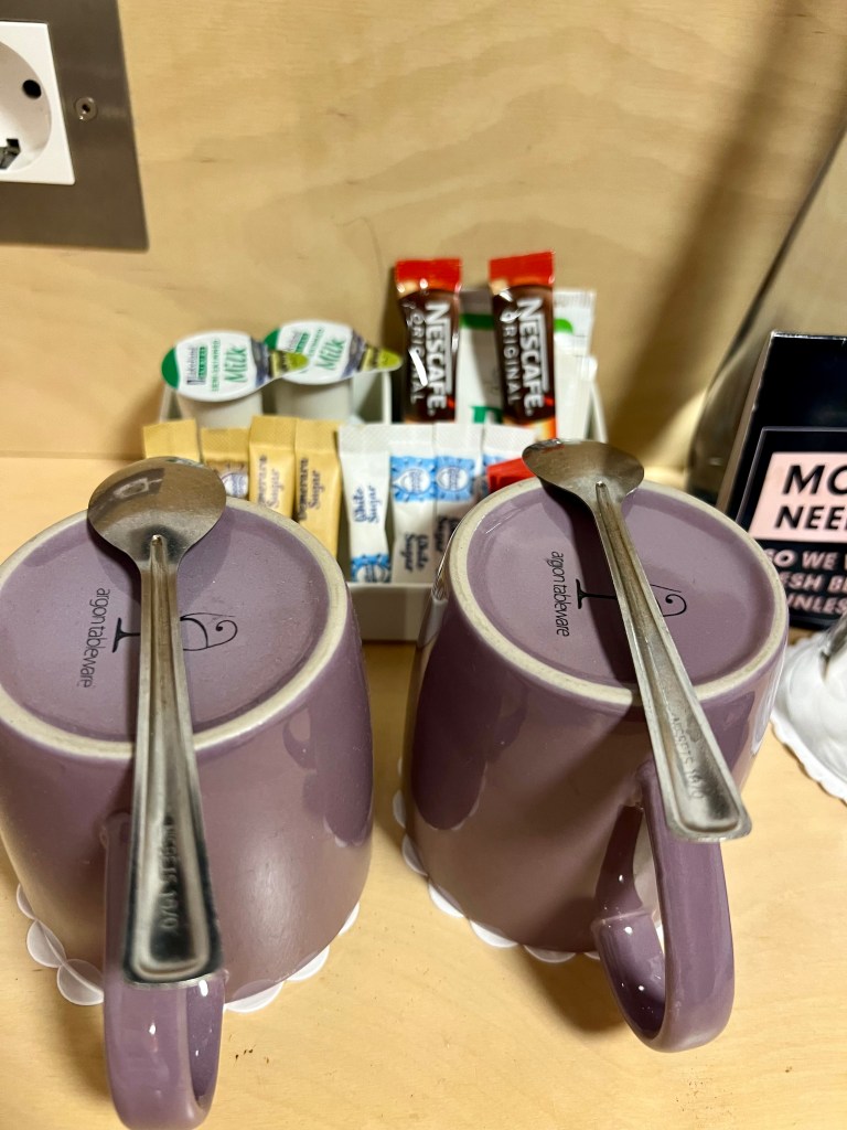 The photo shows a close up of two purple mugs upside down on a light wooden surface. There is a teaspoon on top of each mug. Behind the mugs slightly out of focus is a selection of tea, coffee and sugar sachets. Part of a plug socket can be see on the wall behind.