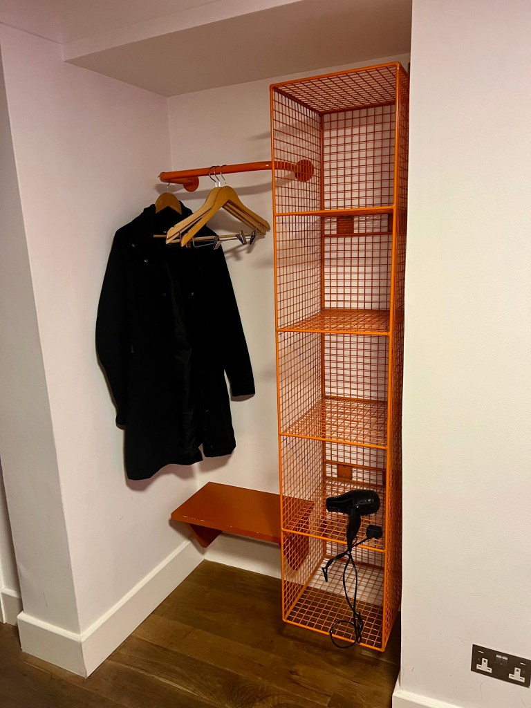 The photo shows an alcove in the bedroom. The walls are white and the floor is dark wood laminate. In the alcove there is an orange metal rack shelf unit with five cube holes and a small orange bench/shelf. Above the bench there is an orange rail withe 3 empty wooden coat hangers and a black knee length coat hanging.