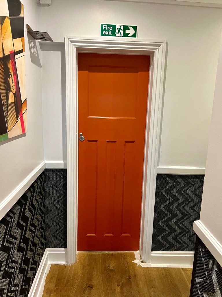 The photo shows a bedroom door painted orange. The walls of the hallway are painted white at the top with a wooden rail at hip height. The bottom of the wall is wallpapered in a black and grey vertical zig zag pattern.