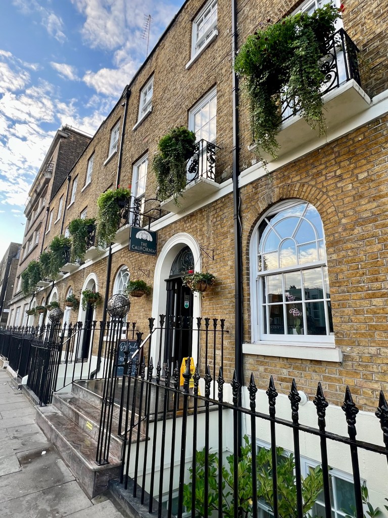 The photo shows the outside of the California Hotel, looking down the street. In the foreground are the black iron railings, behind are the arched ground floor windows and the second floor windows with balconies and greenery.