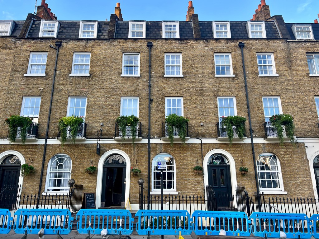 The photo shows a row of 4 brick town houses. Each has a black front door with a white archway and then 3 floors or windows above.
In front of the houses is a room of bright blue barriers.