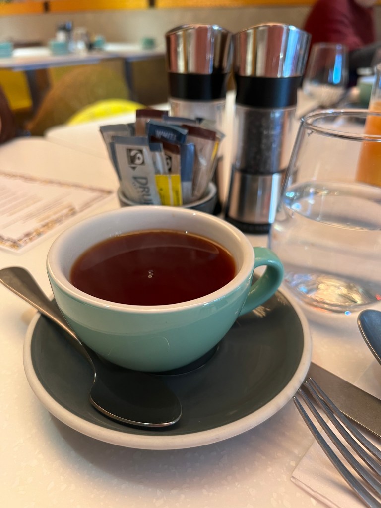 The photo shows a green cup and saucer filled with black coffee, there is a teaspoon on the saucer. Behind the cup are silver salt and pepper mills, a small sugar bowl containing sachets of sugar and a glass of water.