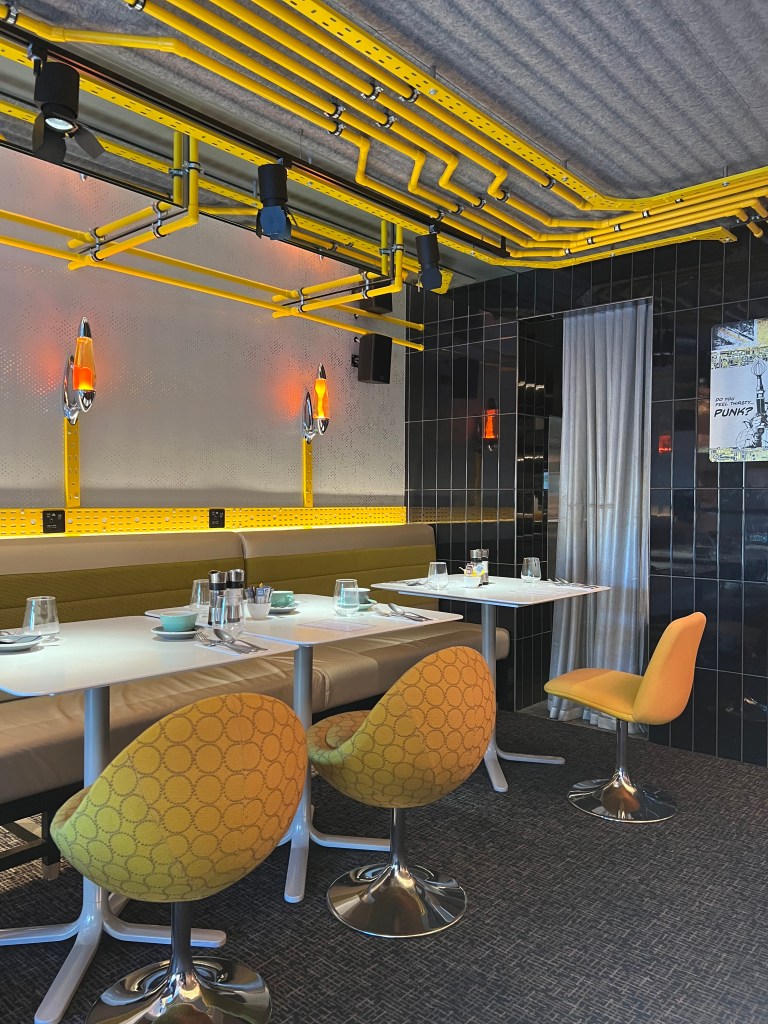 The photo shows two of the tables in the restaurant. the table on the left seats four and has two yellow seats in the foreground and cream banquette seating in the background. The table on the right seats two and has one yellow chair. The wall behind is painted white and has two orange lave lamps on the wall. the ceiling is decorated with bright yellow pipes.