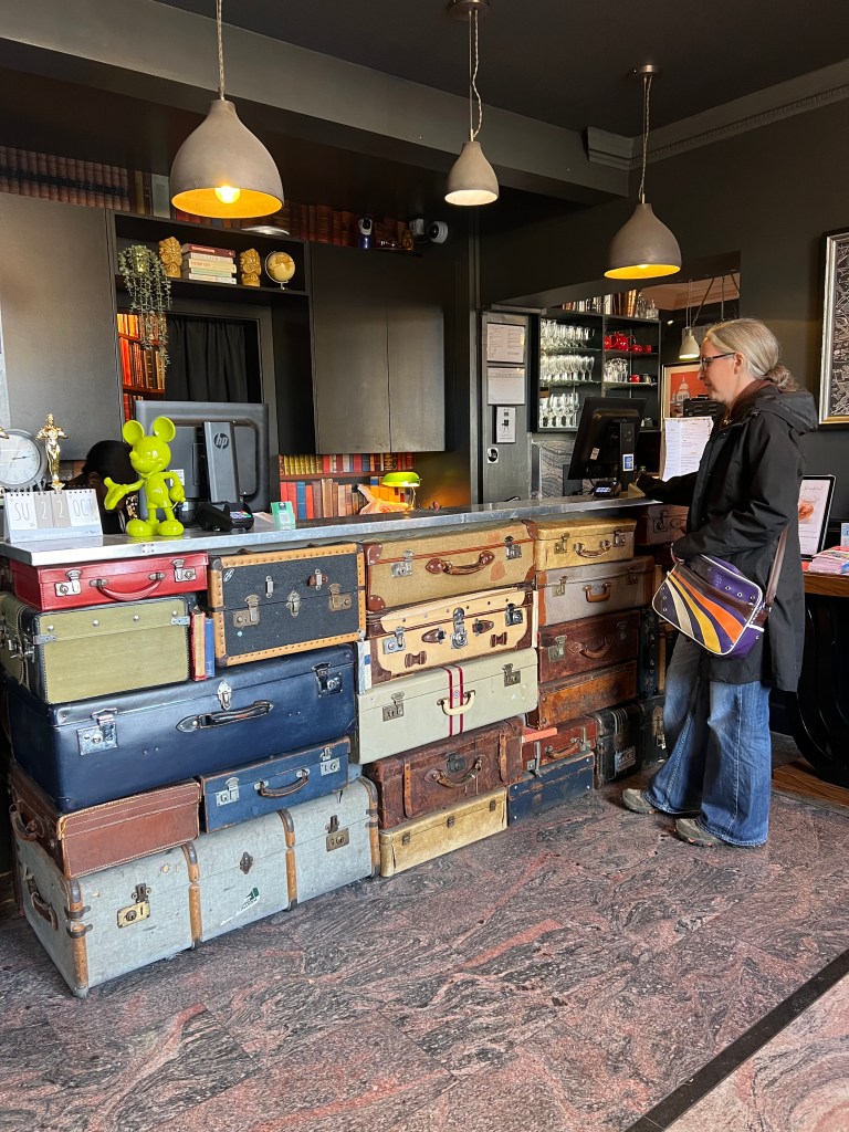 The reception of the hotel which looks like it is balanced on top of a stack of vintage suitcases. 3 pendant lights hang above the reception desk and there is a lime green Mickey Mouse ornament stood on the counter. On the right of the photo is a forty year old woman with grey hair tied back and a black knee length coat and blue jeans. She is wearing a vintage purple cross body bag.