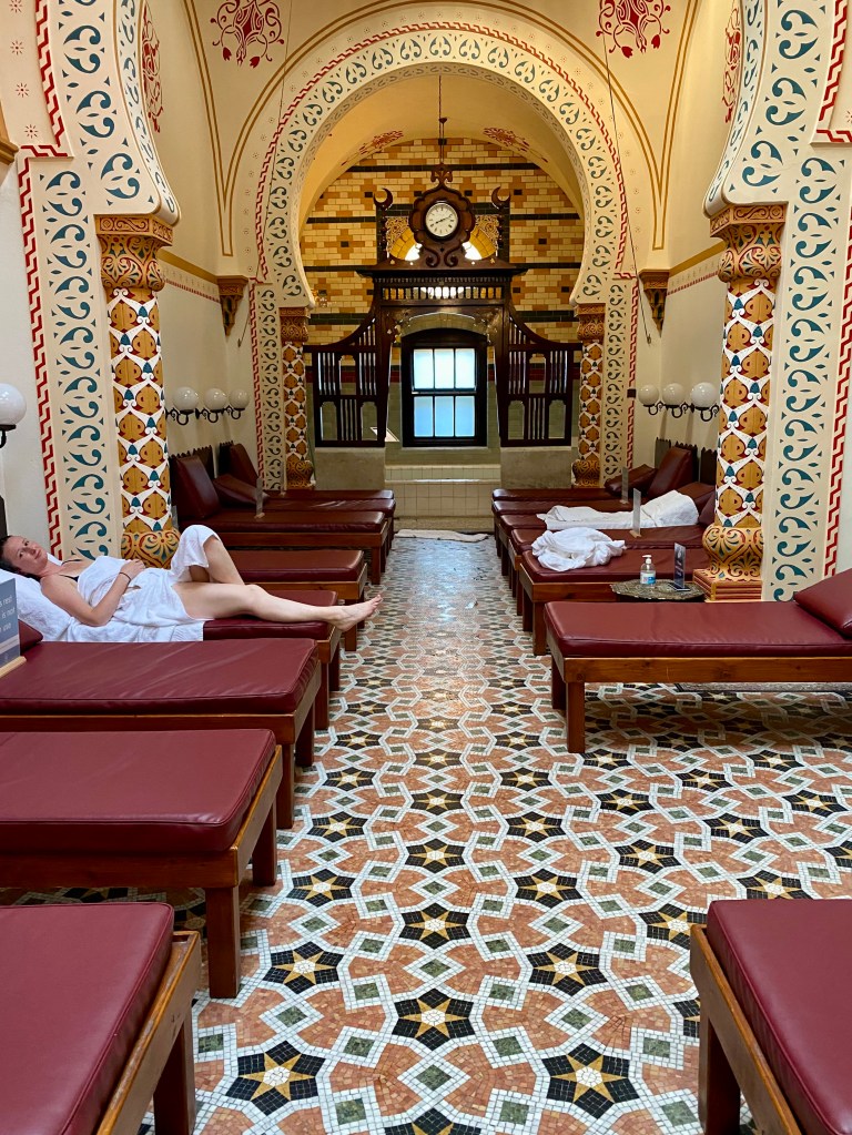 The photo Is looking down the frigidarium room towards the plunge pool. There are several beds down each wall with dark red leather tops. A woman wrapped in a white towel is lying on one of the beds on the left handside. At the end of the room is an archways and a wooden decorative arch that leads to the cold plunge pool.