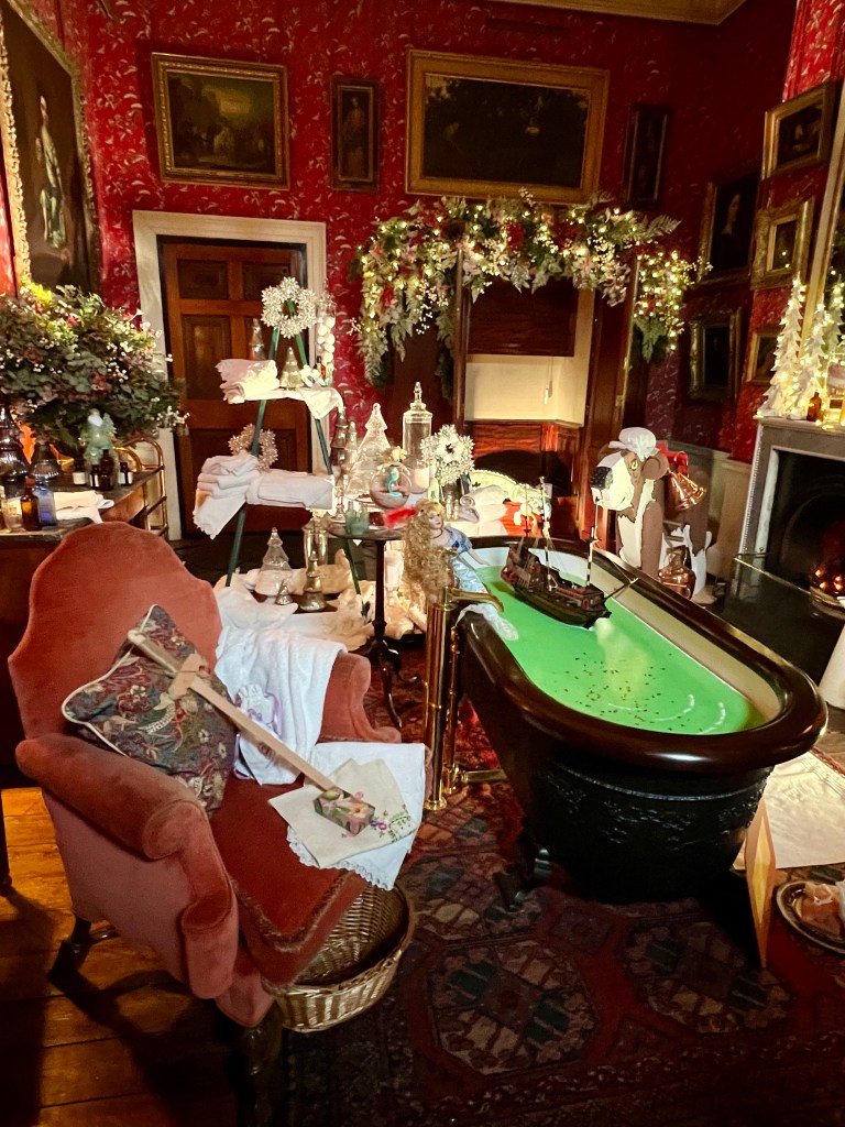 Bath time with Nanna
The room is decorated with dark red wallpaper and gold framed portraits. In the centre of the room is a freestanding bath filled with water. Next to the bath is a large wooden St Bernard dog wearing a maids cap. The room is decorated with garlands and fairy lights. 