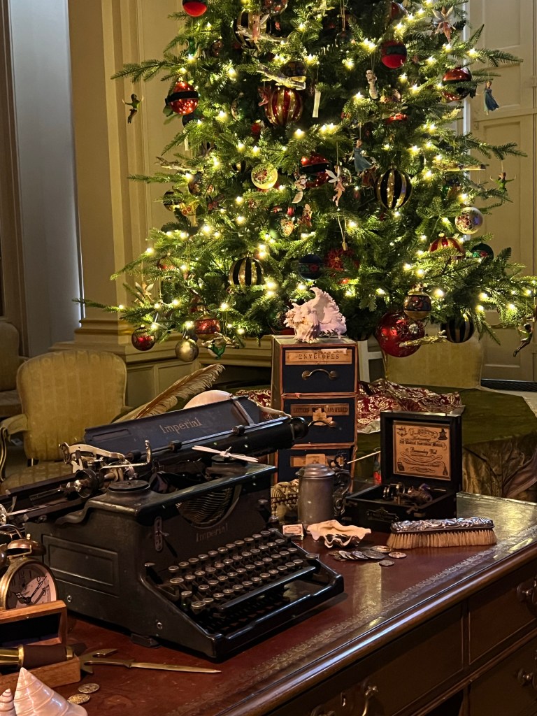 J.M. Barrie's study. A old typewriter sits on a wooden desk with various antique men's items around it. Behind it is a Christmas tree decorated with fairy lights and red baubles. 