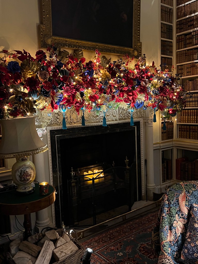 A white stone fireplace with a huge Christmas garland handing above it with red and blue flowers and fairy lights. 