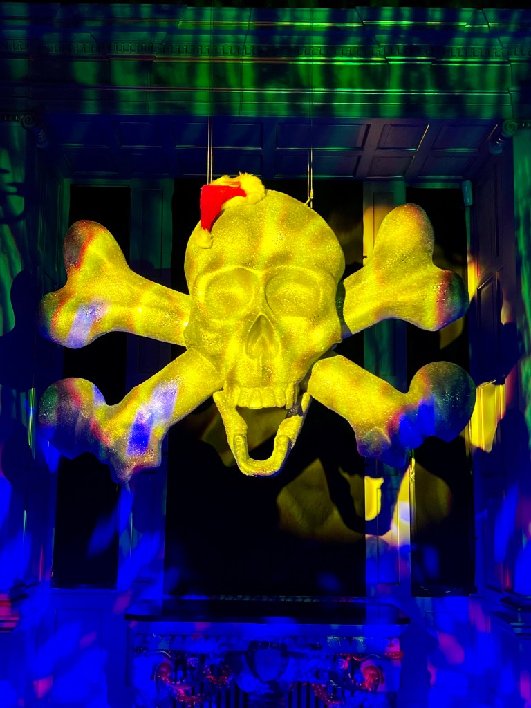 A very large skull and crossbones is hanging from the ceiling and wearing a tiny Santa hat. It is illuminated in yellow in an otherwise dark room. 