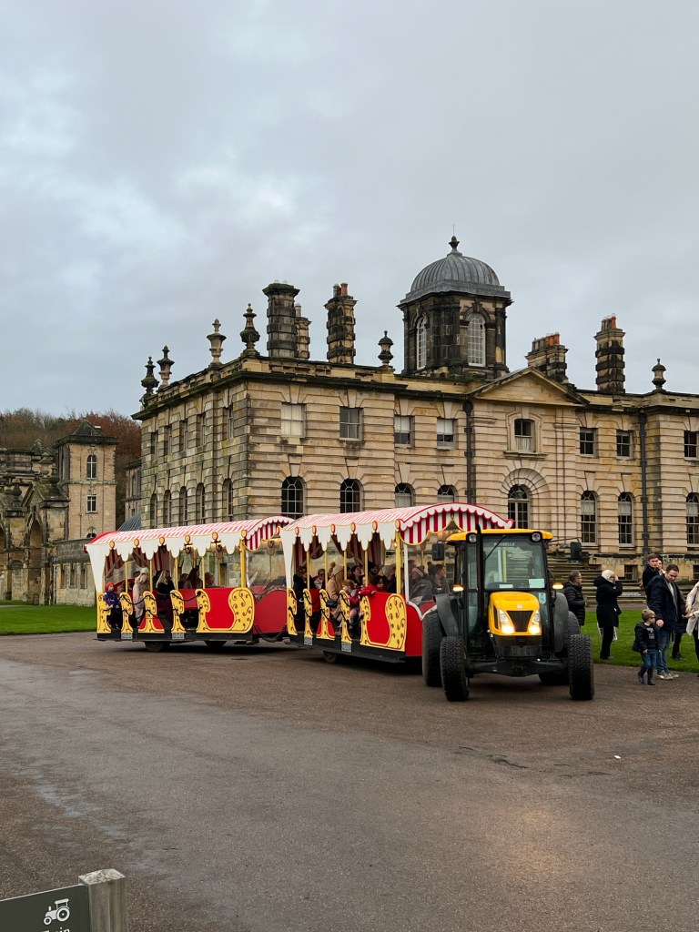 The Kelly Train (Land train) in front of Castle Howard. A yellow tractor pulls two carriages painted in red and yellow. 