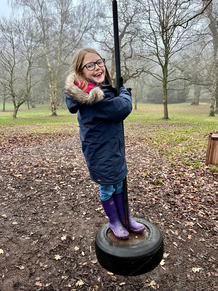 Pip, a 9 year old girl wearing purple wellies and a navy coat with a fur hood stands on a small tire on the end of a black pole. 