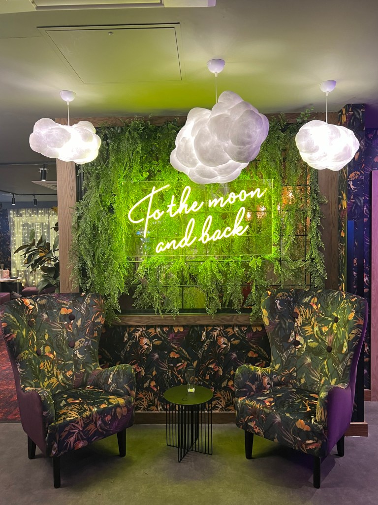 2 green floral high backed arm chairs and a small black side table sit in front of a wall wallpapered with the same pattern as the chairs. The top half of the wall has a neon sign that reads To the moon and back and fake greenery. The lights hangings from the ceiling are shaped like clouds. 