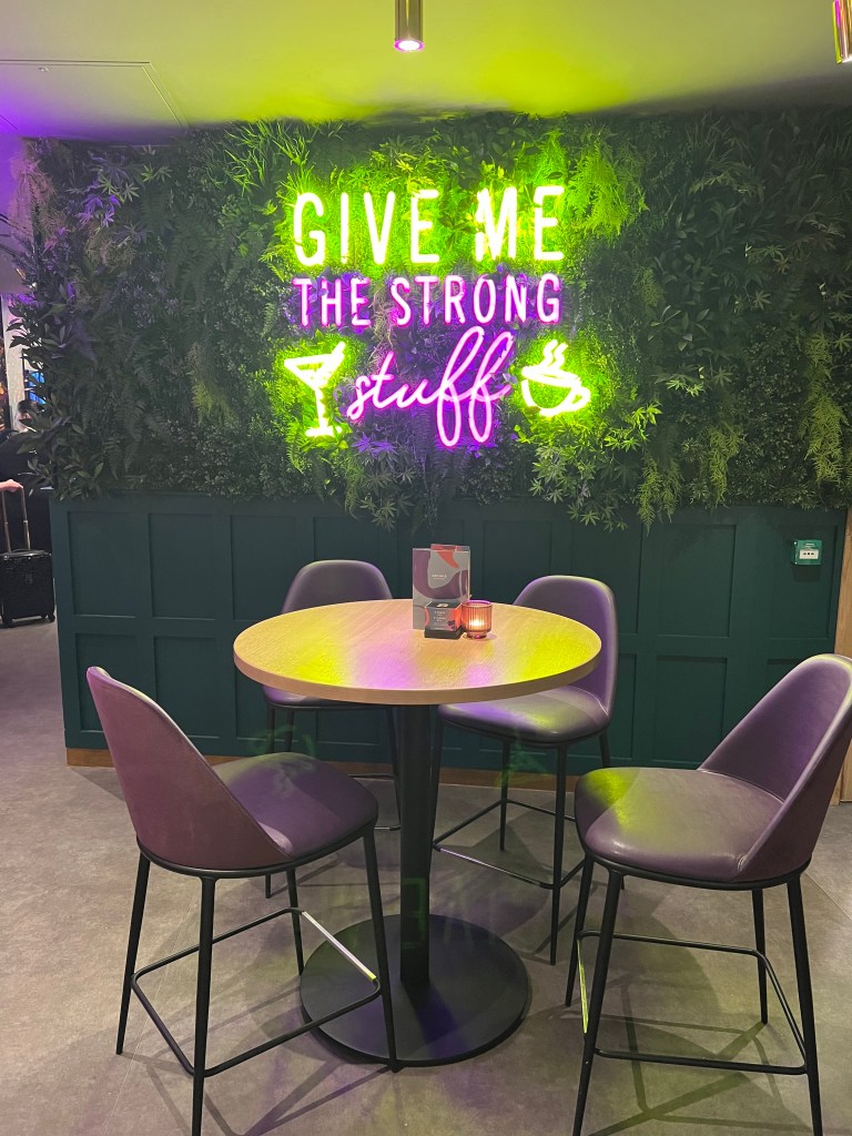 A round bar table and 4 brown leather chairs. The wall behind are painted darl green with more fake greenery. The large neon sign on the wall reads Give me the strong stuff in green and pink. 