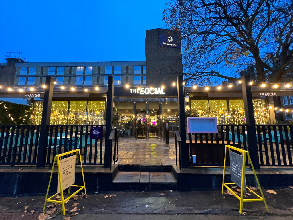 The front of the hotel at night showing the outside area of the Social restaurant decorated in fairy lights with the Premier Inn building behind and a tower with the Premier Inn logo.