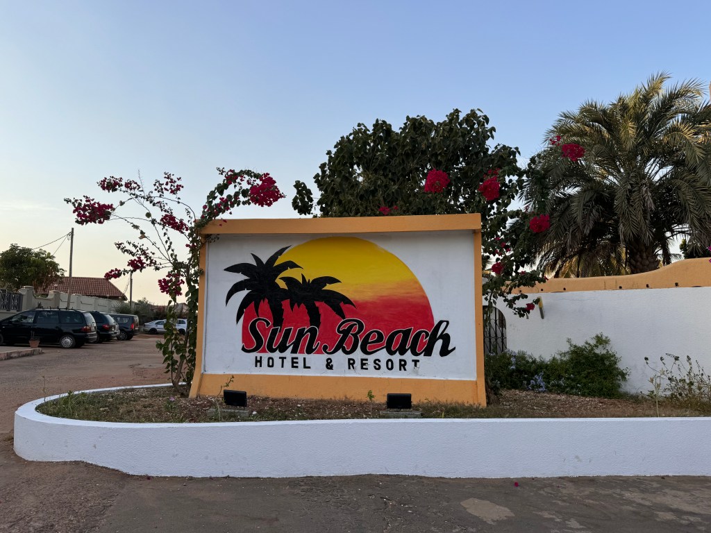 The Sun Beach hotel sign. A large concrete sign with the words Sun Beach Hotel & Resort, behind the words are a semi circle painted in red and yellow like a sun set and the silhouette of two palm trees. the sign is set in a flower bed with trees behind it. 