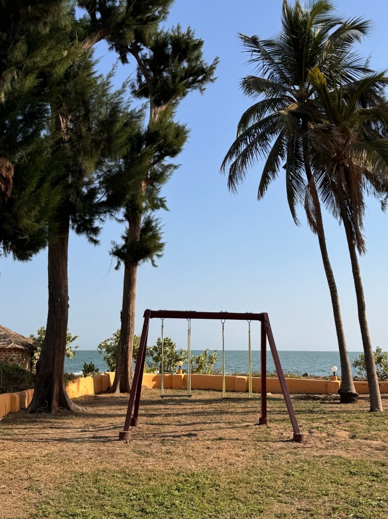 A double wooden swing with the sea behind and palm trees on either side.