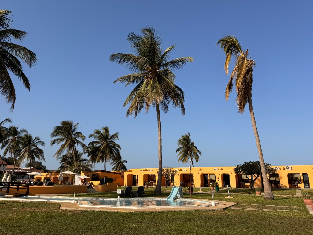 The children's splash pool with a small toddler size slide in to the pool. Behind is a low yellow building and palm trees.