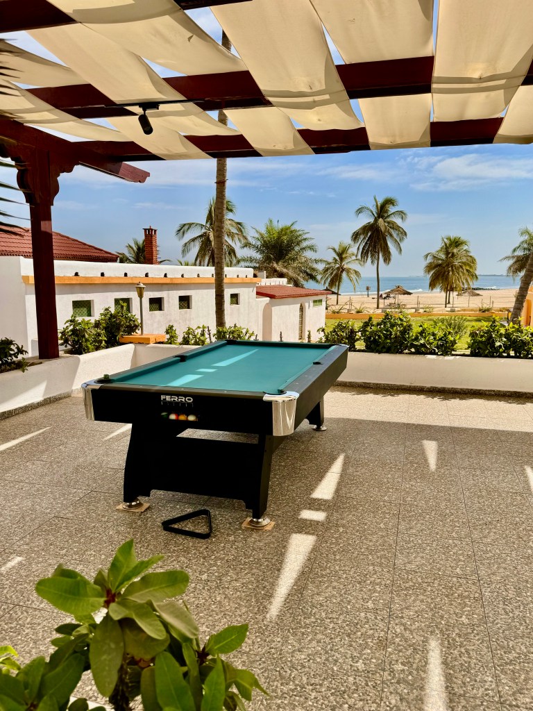 The pool table on a stone patio with a roof canopy over it for shade. behind you can see a white building, the beach and palm trees. 