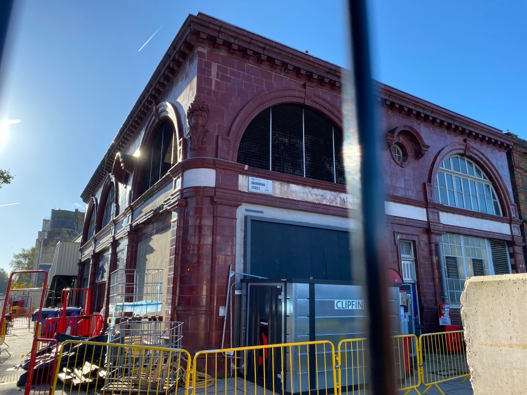 The outside of an original underground station taken from between the fence railings. The outside of the building is covered in oc blood red tiles and has various construction material and fencing outside.