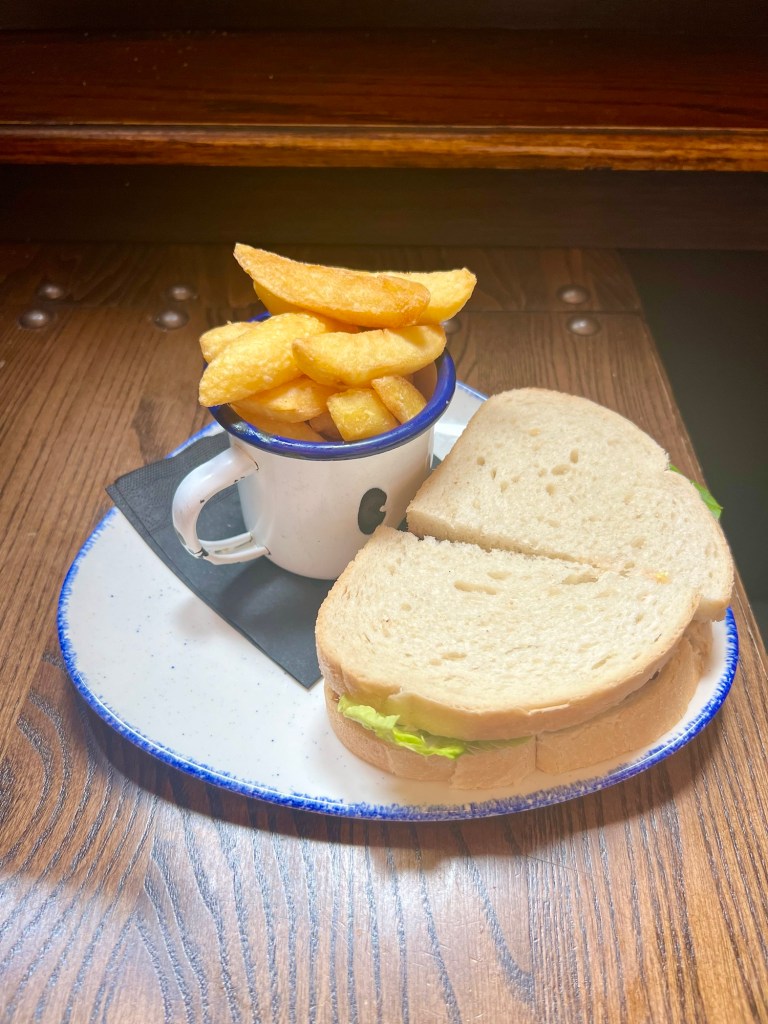 A fishmonger sandwich on white bread with an enamel mug full of chips. 