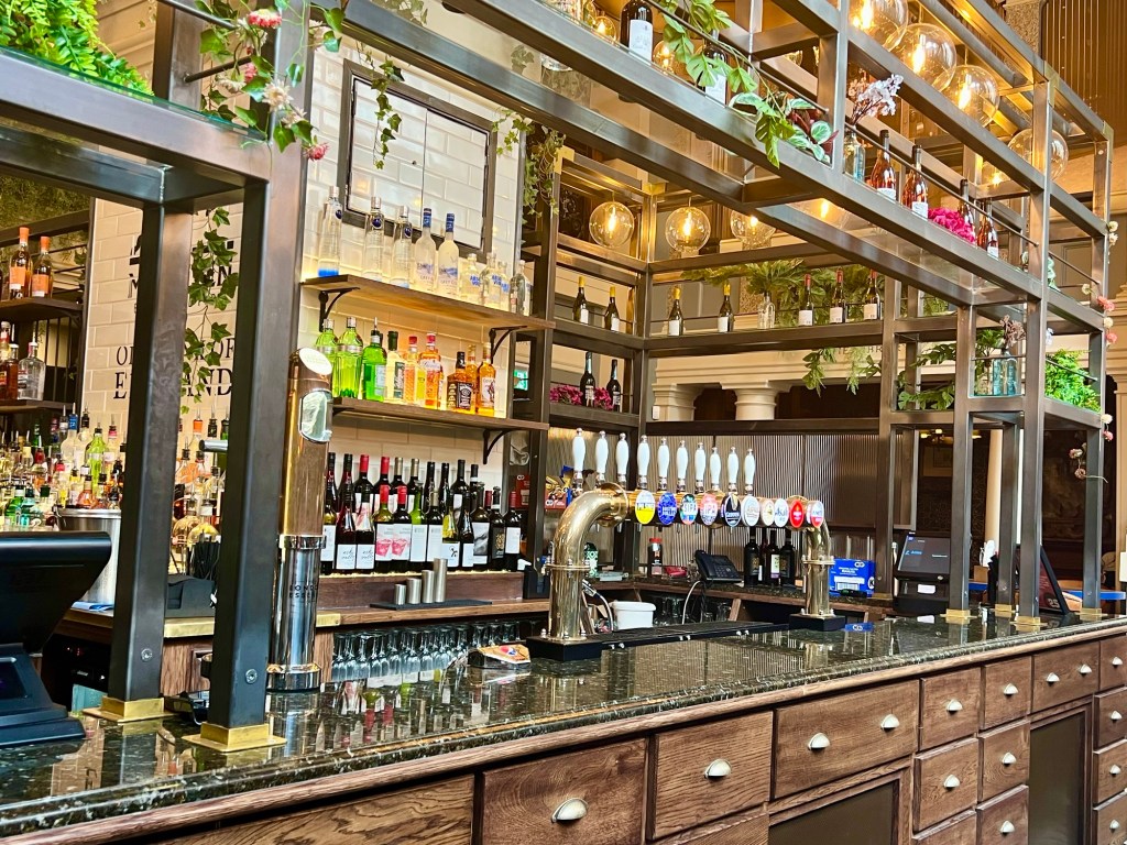 The bar of the pub. The counter looks like an old shop counter with wooden drawers and a marble top. On the wall behind are bottles of wine and spirits. 
