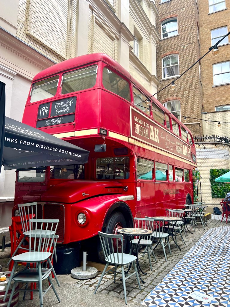 A bright red route master double decker bus parked in a courtyard surrounded by buildings. The floor has blue and brown geometric tiles and there and several small tables and chairs next to the bus. 