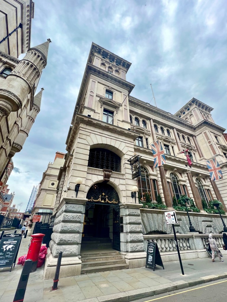 The outside of the building. An imposing 4 storey building with arched windows, columns, white stone and marble. There are 2 Union Jacks hanging from the front of the building and an arched entrance with black wrought iron gates. 