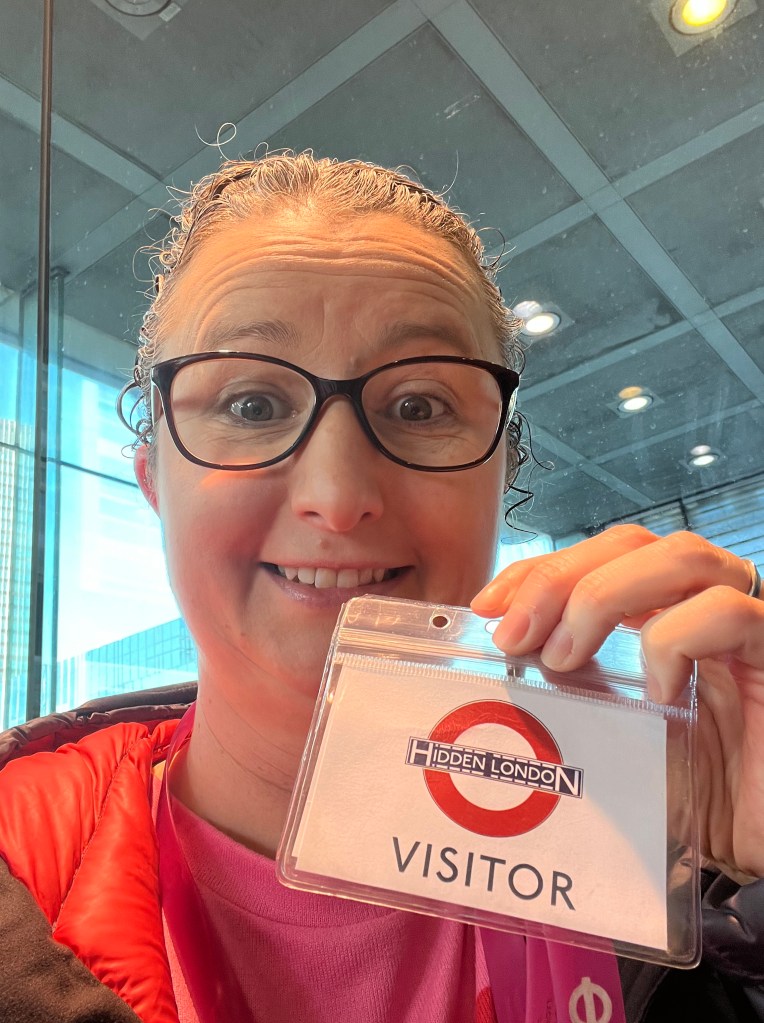 A white woman in her forties wearing a bright pink top and a navy puff jacket with a red lining. She has her hair tied back and is wearing glasses. She is holding up a visitor badge on a lanyard which reads Visitor and has the Hidden London underground logo, looking at the camera and pulling a surprised face.
