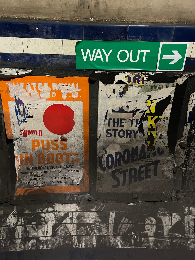 Two damaged and dirty posters on the wall of the tunnel. the right one is for Coronation Street and the left for Puss in Boots. There is a Green way out sign on the wall above the posters.