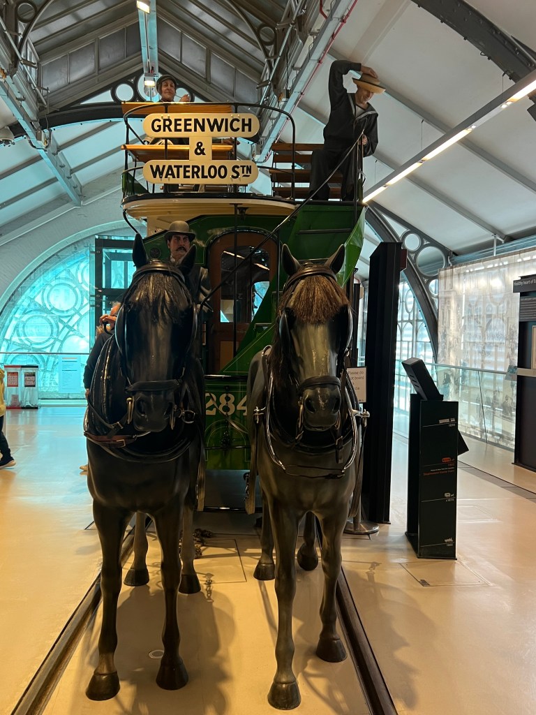 An original horse drawn green bus pulled by two model horses. The driver manikin sits on the open top holding the reins. 