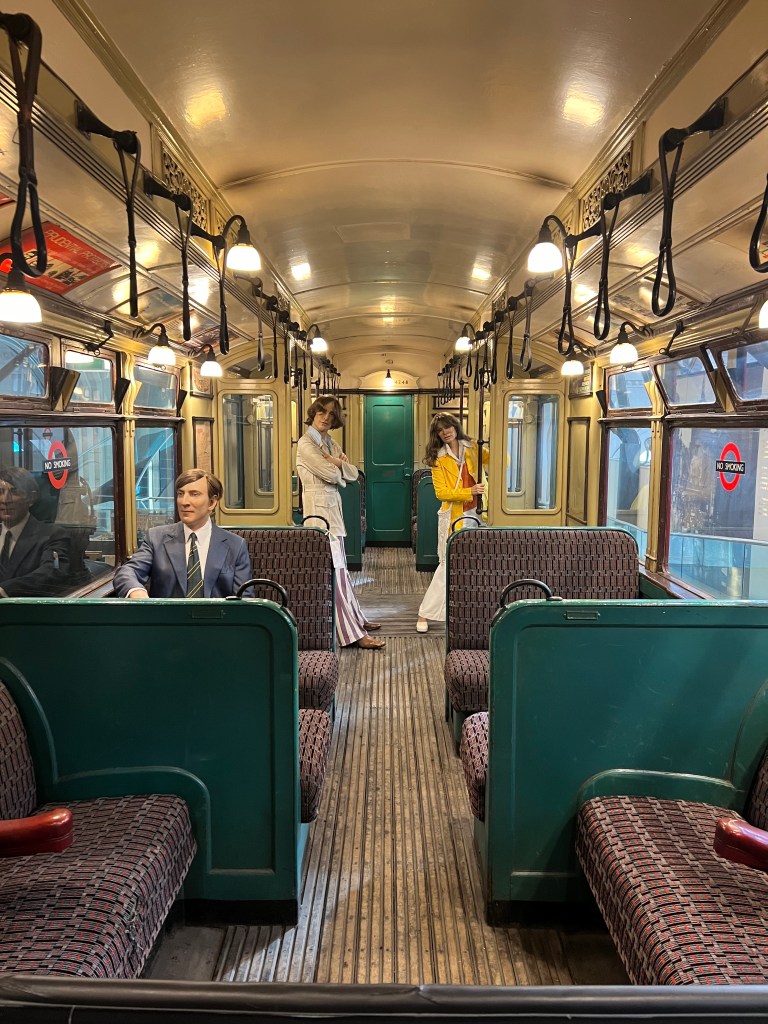 A view down an older underground carriage. There are 3 manikins wearing 1970s style clothing stood and sat in the carriage. The carriage is painted in cream and dark green and upholstered in red and green. 