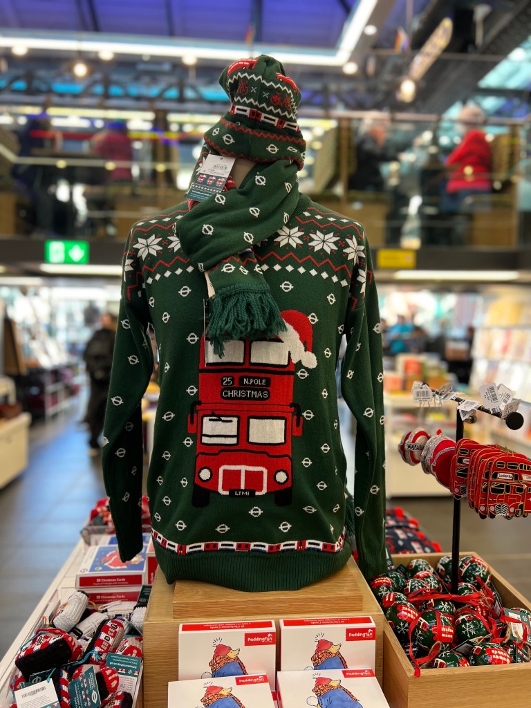 A Christmas jumper on display in the shop. The jumper is dark green and has a red double decker bus on the front and white snowflakes. The jumper has a matching scarf and hat with it. 