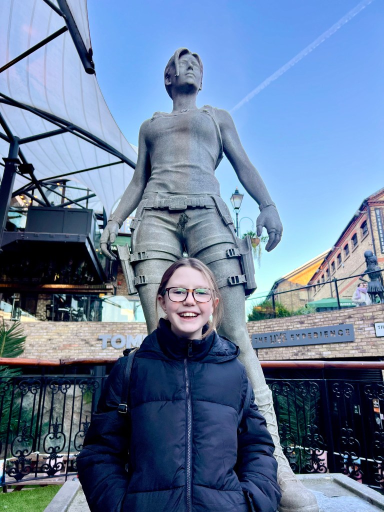 An 11 year old girl wearing a black puffer jacket and glasses is smiling at the camera. She is standing in front of a large statue of Lara Croft. 