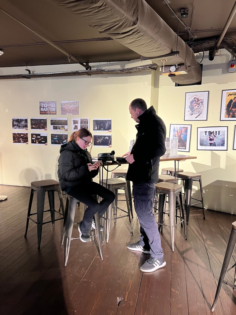 An 11 year old girl, wearing black is sat on a bar stool, next to her is a man also wearing black. They are both looking at something in her hands. 