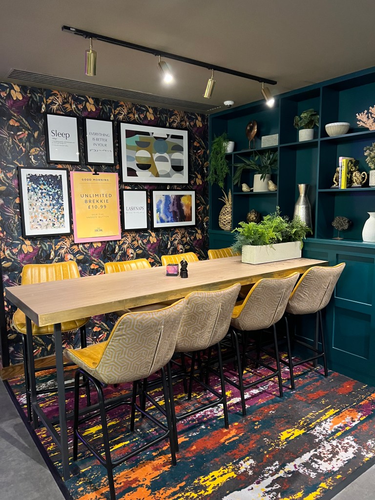 A seating area with a long bar table and 8 high bar chairs. The wall behind is papered in a dark green floral paper and there isa shelving unit on the other wall painted in dark green.