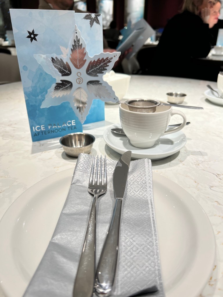 Afternoon tea place setting. A white plate with a silver paper napkin and knife and fork, behind is a white cup and saucer with a tea strainer in it. Stood up behind it is a blue menu with a silver snowflake on the front.