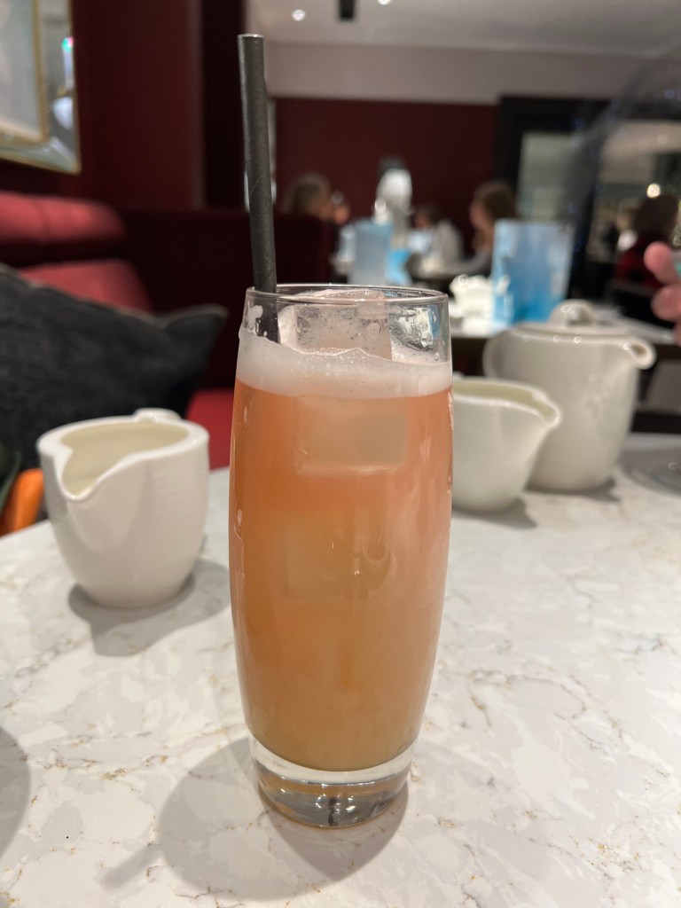 A photo of a pink mocktail in a tall glass stood on a white table cloth. Behind can be seen a white teapot and milk jug.