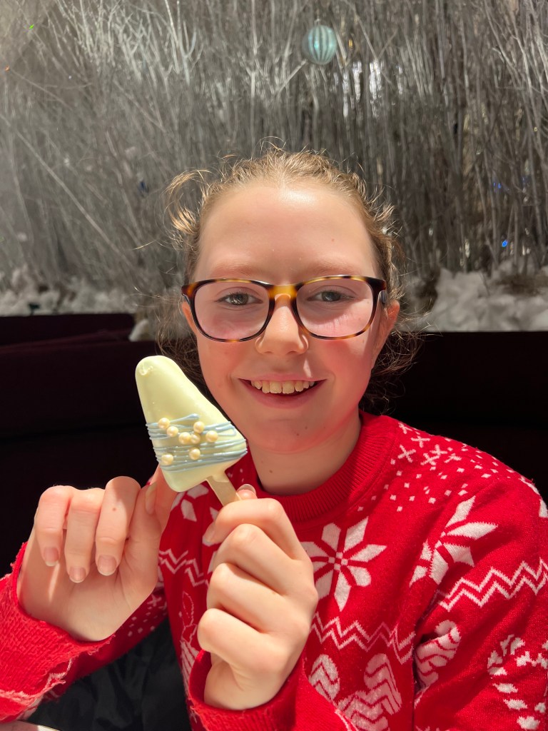 An 11 year old girl with her hair tied back and tortoise shell glasses is wearing a red and white Christmas jumper. She is holding a triangular shaped cake pop decorated with white chocolate and blue icing.