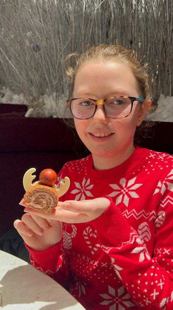 An 11 year old girl with her hair tied back and tortoise shell glasses is wearing a red and white Christmas jumper. She is holding a slice of chocolate Swiss roll with white chocolate antlers and a red chocolate ball.