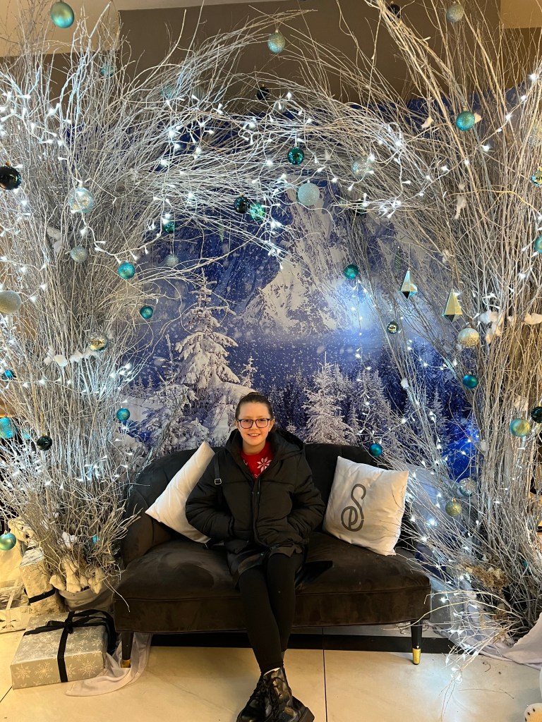 An 11 year old girl with her hair tied back and tortoise shell glasses is wearing a red and white Christmas jumper and a black coat. She is sitting on a small sofa under an archway of white branches and fairy lights.
