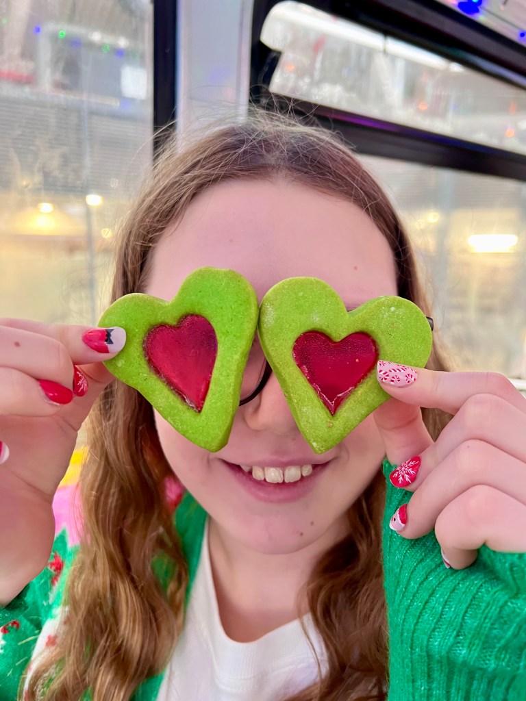 An 11 year old girl wearing a brightly coloured green and pink Christmas cardigan, her hair down and glasses. She is holding up two green heart shaped biscuits with red centres in front of her eyes. 
