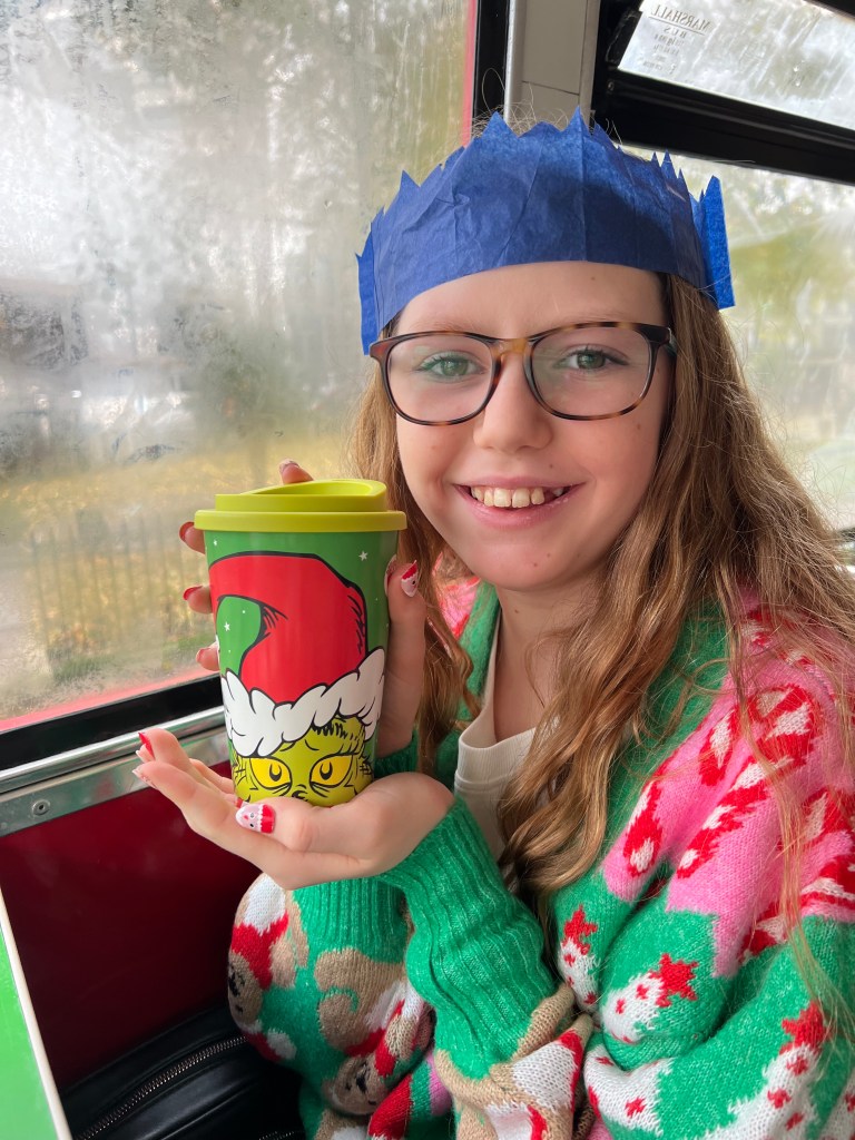 An 11 year old girl wearing a brightly coloured green and pink Christmas cardigan, her hair down and glasses. She is wearing a blue Christmas cracker crown and holding a reusable cup with the grinch's face on it. 