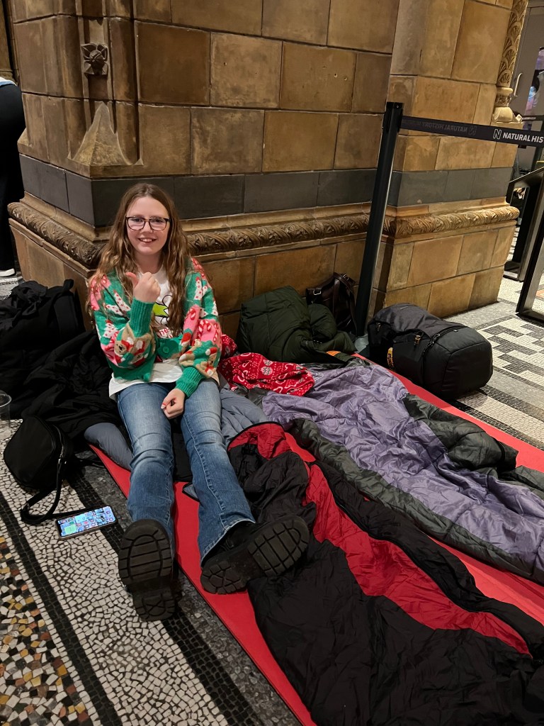 A white 11 year old girl wearing blue jeans and a colourful sweater is sat on a a camping mat and sleeping bag which is laid on a tiled floor next to a stone wall.