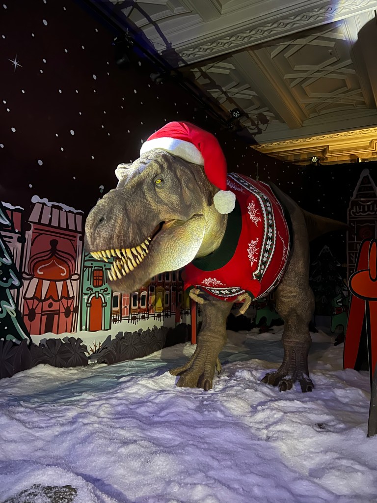 A large animatronic T-Rex wearing a red and green Christmas jumper and a Santa hat.