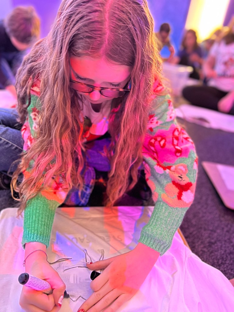 A white 11 year old girl wearing blue jeans and a brightly coloured Christmas jumper is sat on the floor drawing on a t-shirt with a black marker pen.